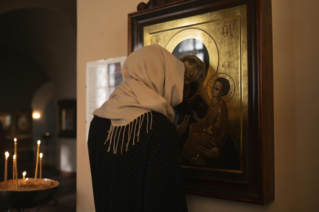 A woman with a headscarf prays before an icon in a dimly lit church, creating a serene religious atmosphere.