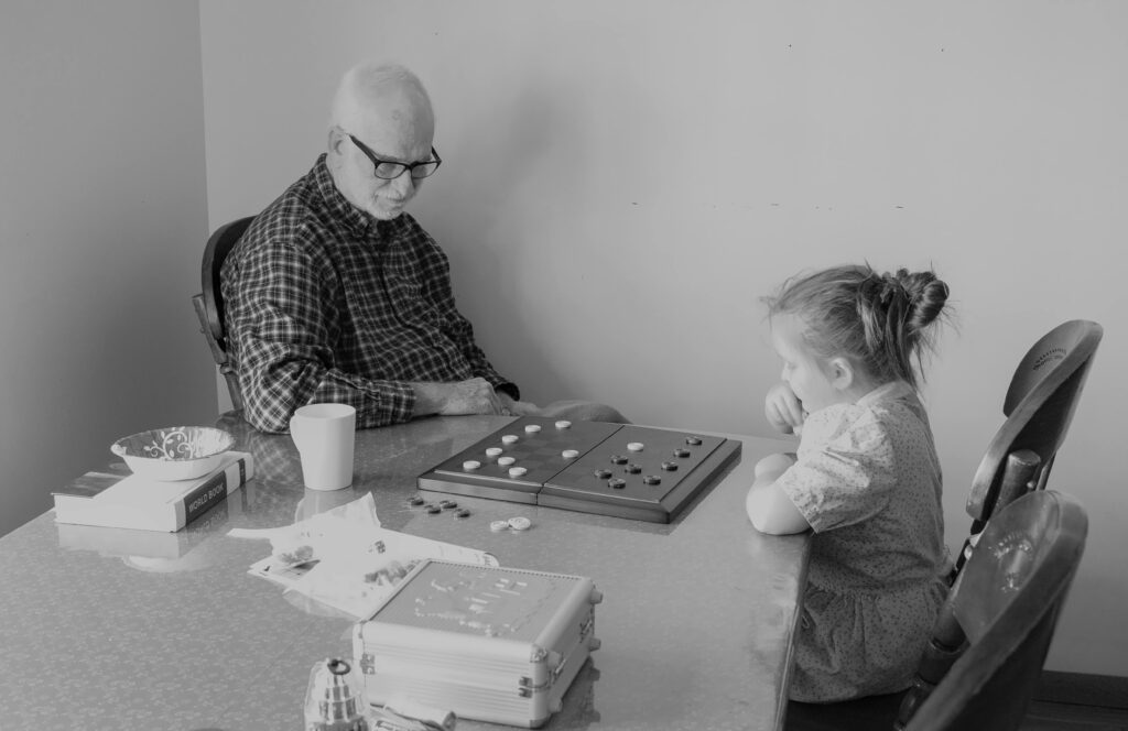 Black and white photo of grandparent with grandchild playing a board game indoors.